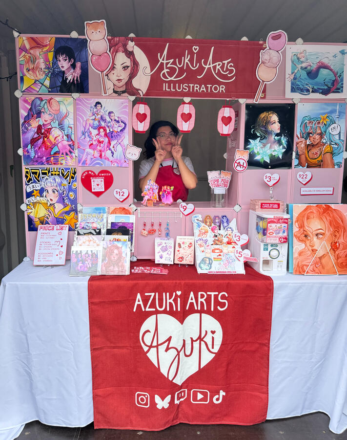 Azuki Arts artist alley vendor booth at KW Creators Illustrators Bazaar 2025 featuring anime and gaming prints, stickers, keychains, and pins.